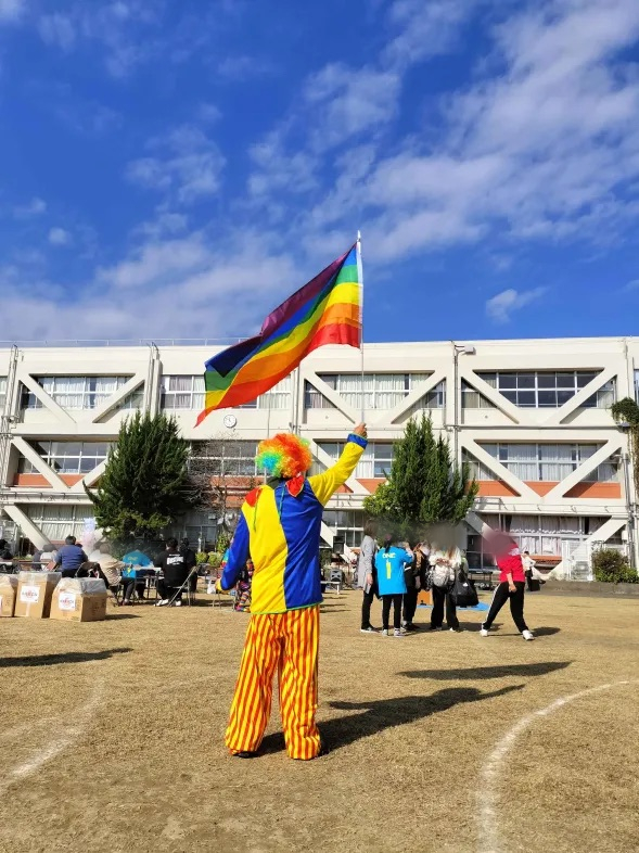 清瀬七小の校舎を背景に、虹色の服を着た人がレインボーフラッグを掲げている写真